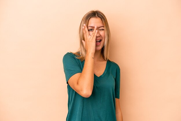 La giovane donna caucasica isolata su fondo beige lampeggia alla macchina fotografica attraverso le dita, imbarazzata che copre il viso.