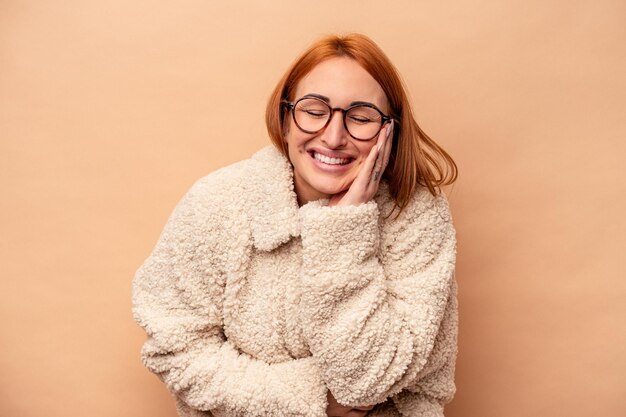 La giovane donna caucasica isolata su fondo beige ride felice e si diverte a tenere le mani sullo stomaco