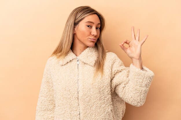 La giovane donna caucasica isolata su fondo beige strizza l'occhio e tiene un gesto ok con la mano.