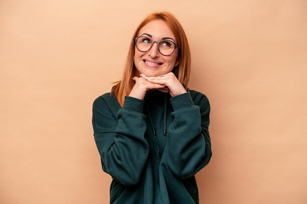 La giovane donna caucasica isolata su fondo beige tiene le mani sotto il mento, sta guardando felicemente da parte.