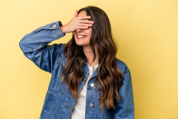 La giovane donna caucasica isolata su fondo giallo copre gli occhi con le mani, sorride ampiamente in attesa di una sorpresa.