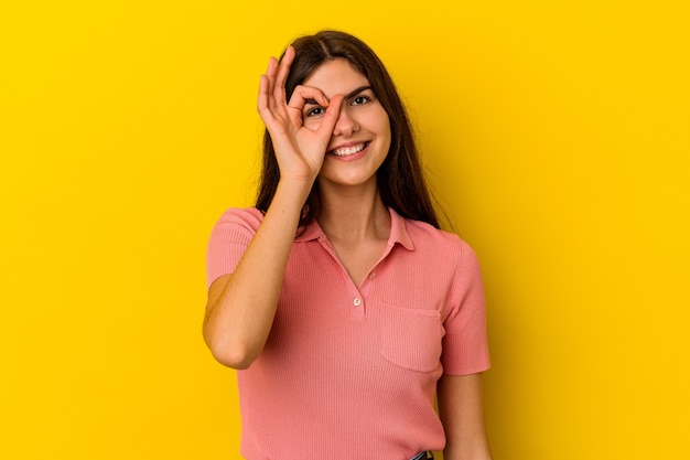 La giovane donna caucasica isolata su fondo giallo ha eccitato mantenendo il gesto giusto sull'occhio.