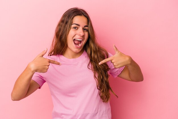 La giovane donna caucasica isolata su fondo rosa ha sorpreso indicando con il dito, sorridendo ampiamente.