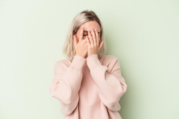 La giovane donna caucasica isolata su fondo verde sbatte le palpebre alla macchina fotografica attraverso le dita, imbarazzata che copre il viso.