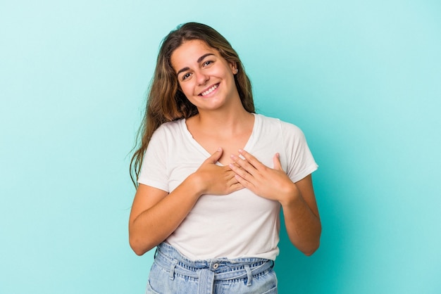 La giovane donna caucasica isolata su sfondo blu ha un'espressione amichevole, premendo il palmo sul petto. Concetto di amore.