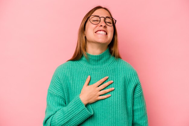 La giovane donna caucasica isolata su sfondo rosa ride ad alta voce tenendo la mano sul petto