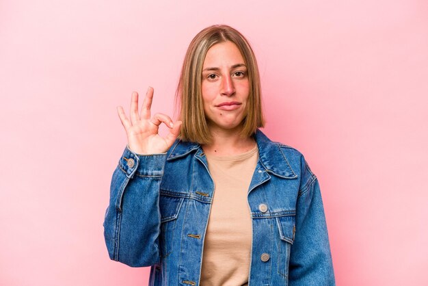 La giovane donna caucasica isolata su sfondo rosa strizza l'occhio e tiene un gesto giusto con la mano