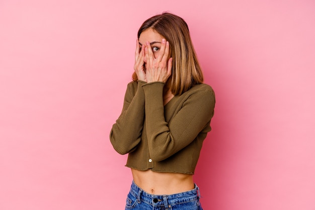 La giovane donna caucasica isolata sul muro rosa sbatte le palpebre attraverso le dita spaventata e nervosa