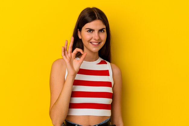La giovane donna caucasica isolata sulla parete gialla strizza l'occhio e tiene un gesto giusto con la mano