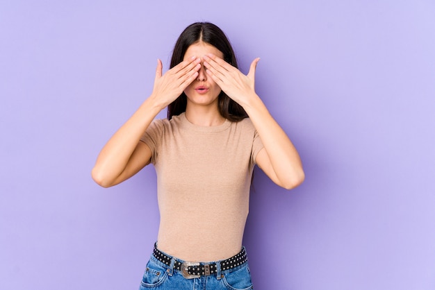 La giovane donna caucasica isolata sulla parete viola ha paura degli occhi di copertura con le mani.