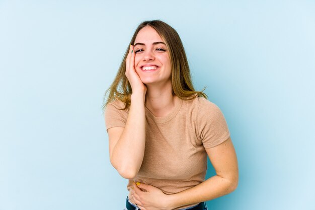 La giovane donna caucasica isolata sullo spazio blu ride felicemente e si diverte a tenere le mani sullo stomaco.