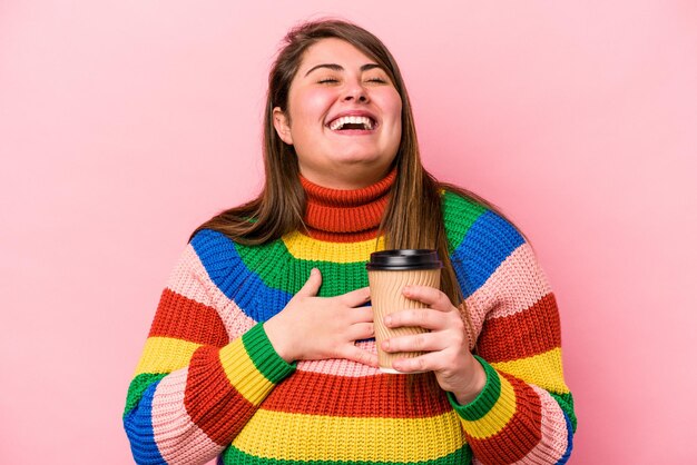 La giovane donna caucasica sovrappeso che tiene il caffè da asporto isolato su sfondo rosa ride ad alta voce tenendo la mano sul petto.