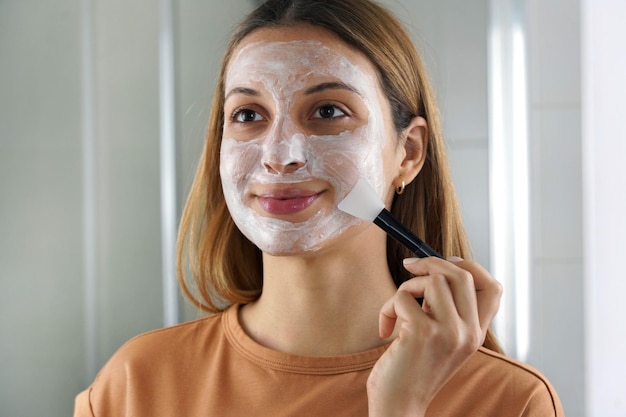 La giovane donna che applica una maschera idratante con una spazzola per il viso si guarda allo specchio a casa