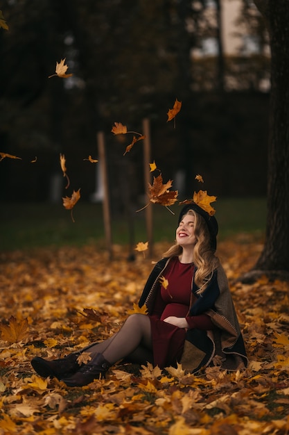 La giovane donna che indossa un vestito rosso e un cappello nero si siede a terra sotto le foglie gialle che cadono su ...