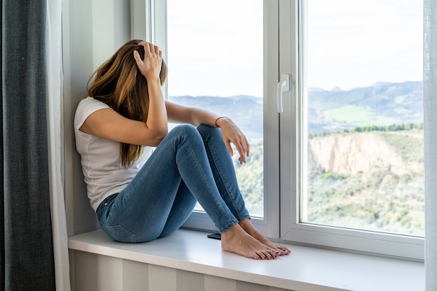 La giovane donna che si siede nella finestra della sua casa guarda fuori. Concetto di stare a casa.