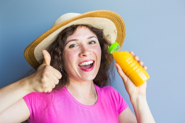 La giovane donna che tiene una bottiglia con succo e che mostra i pollici aumenta il gesto. Cibo sano, disintossicazione e concetto di dieta