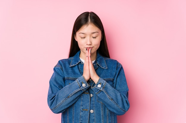 La giovane donna cinese che posa in una parete rosa isolata tenendo le mani dentro prega vicino alla bocca, si sente sicura.