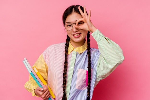 La giovane donna cinese dell'allievo che tiene i libri che portano una camicia e una treccia multicolori di modo, isolate su fondo rosa ha eccitato mantenendo il gesto giusto sull'occhio.