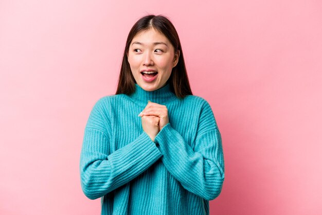 La giovane donna cinese isolata su sfondo rosa tiene le mani sotto il mento sta guardando felicemente da parte