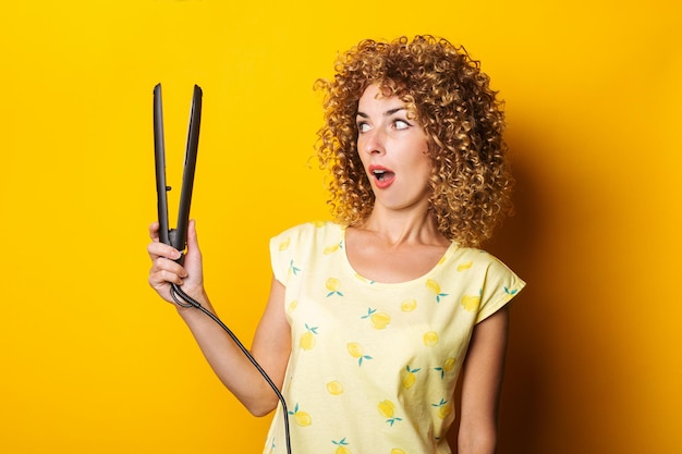 La giovane donna con i capelli ricci guarda sorpresa la piastra per capelli su uno sfondo giallo.