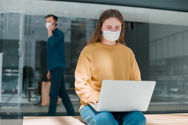 La giovane donna con una maschera protettiva usa il suo laptop per strada. concetto di tutela della salute