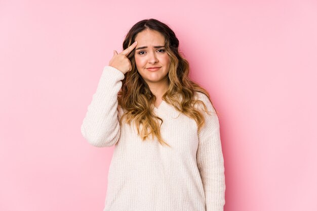 La giovane donna curvy che posa in una parete rosa ha isolato la mostra del gesto di delusione con l'indice.