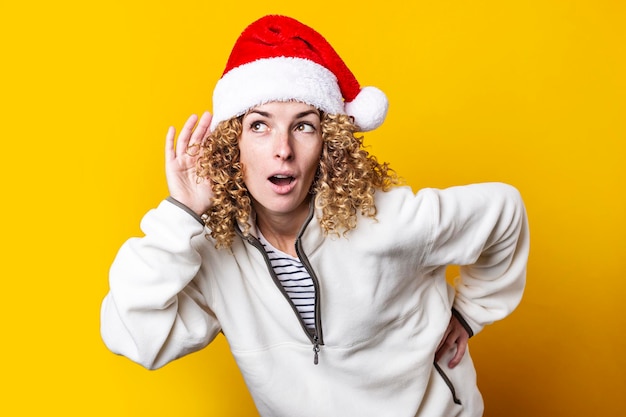 La giovane donna dai capelli ricci in cappello di babbo natale origlia su uno sfondo giallo.