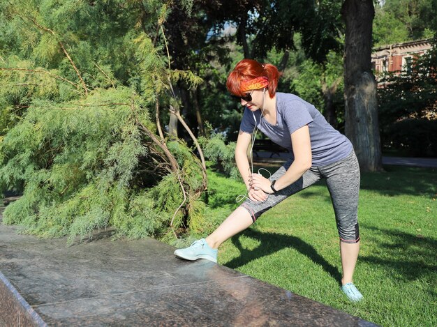 La giovane donna dai capelli rossi in abiti sportivi ascolta con le cuffie e fa esercizi da sola nel parco