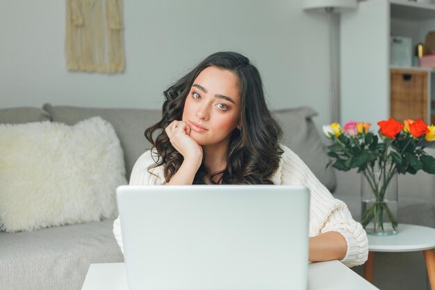 La giovane donna dai capelli scuri lavora online utilizzando il laptop Bellissimo modello in maglione lavorato a maglia in ufficio a casa Casa accogliente Freelans