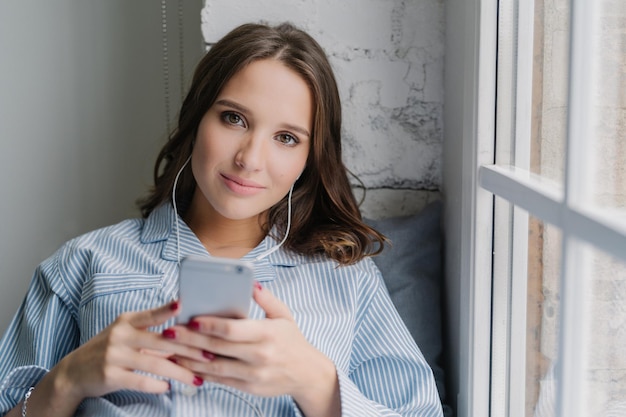 La giovane donna dall'aspetto piacevole soddisfatta del buon suono negli auricolari tiene il telefono cellulare moderno vestito casualmente gode del tempo libero si siede sul davanzale della finestra La femmina ascolta le canzoni della playlist indoor