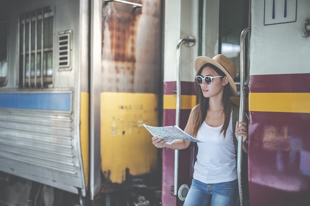 La giovane donna dei viaggiatori con lo zaino che guarda tiene una mappa alla stazione ferroviaria. Giorno del turismo