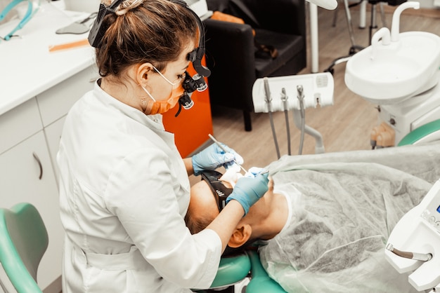 La giovane donna del dentista tratta un paziente un uomo. Il medico usa guanti monouso, una maschera e un cappello. Il dentista lavora nella bocca del paziente, utilizza uno strumento professionale