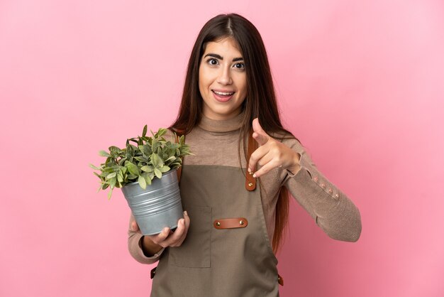 La giovane donna del giardiniere che tiene una pianta isolata sulla parete rosa ha sorpreso e che indica davanti