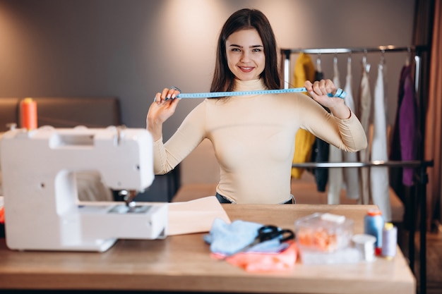La giovane donna del sarto da donna cuce i vestiti sulla macchina da cucire. Tenendo il metro a nastro in officina. Creazione di corsi di design di abbigliamento online.