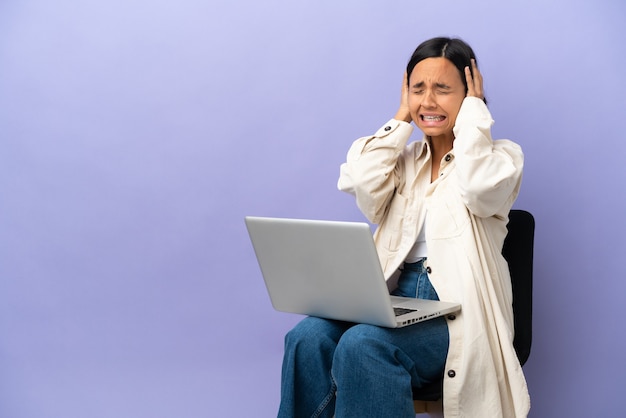 La giovane donna della corsa mista che si siede su una sedia con il laptop isolata sulla parete viola ha sottolineato sopraffatta