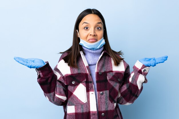 La giovane donna della corsa mista del brunette che protegge con una mascherina e guanti sopra la parete blu che fa i dubbi gesticola