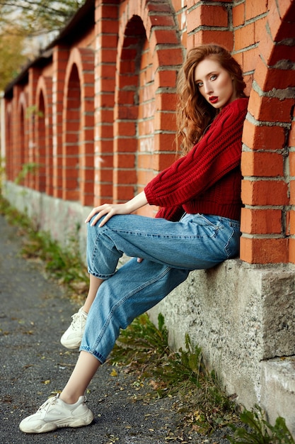 La giovane donna della testarossa in un maglione rosso cammina nel parco. Ritratto di bellezza autunnale di una donna dai capelli rossi alla moda al tramonto