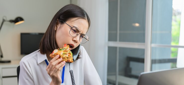 La giovane donna di affari asiatica che mangia la pizza fa uno spuntino mentre parla il telefono con i clienti a causa del lavoro in ritardo.