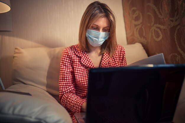 La giovane donna di affari in una mascherina protettiva medica lavora da casa al computer durante l'autoisolamento e la quarantena.