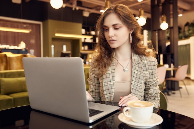 La giovane donna di affari nell'abbigliamento casual astuto beve il caffè e lavora al computer portatile