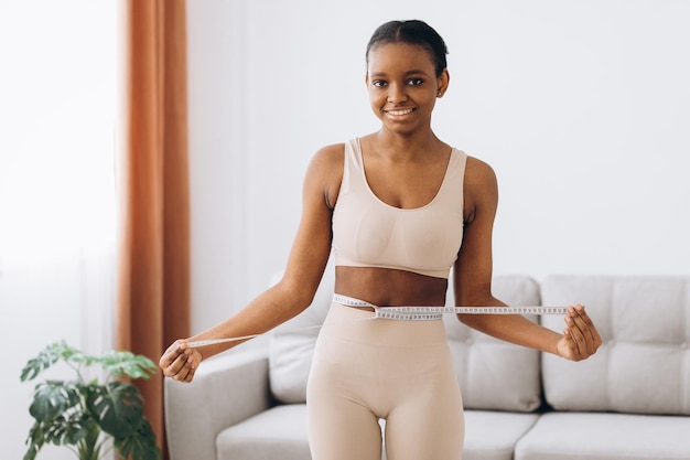 La giovane donna di colore misura la sua vita sullo sfondo del soggiorno. Il concetto di dieta e perdita di peso.