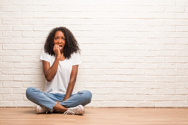 La giovane donna di colore seduta su un pavimento di legno che mangia le unghie, nervosa e molto ansiosa e spaventata per il futuro, prova panico e stress