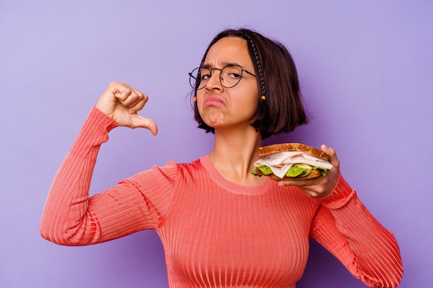 La giovane donna di razza mista che tiene in mano un panino isolato su sfondo viola si sente orgogliosa e sicura di sé, esempio da seguire.