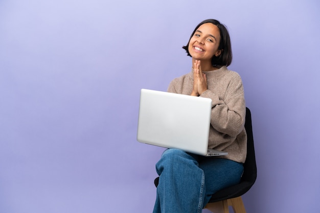 La giovane donna di razza mista seduta su una sedia con il laptop isolato su sfondo viola tiene insieme il palmo. La persona chiede qualcosa