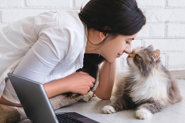 La giovane donna e il suo adorabile gatto si strofinano il naso a vicenda. Il soffice animale domestico di famiglia giace sul tavolo di lavoro con il laptop nelle vicinanze