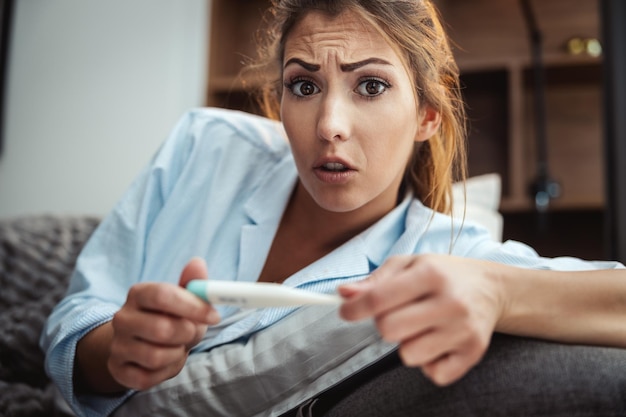 La giovane donna è malata sul divano di casa e sta guardando il termometro.