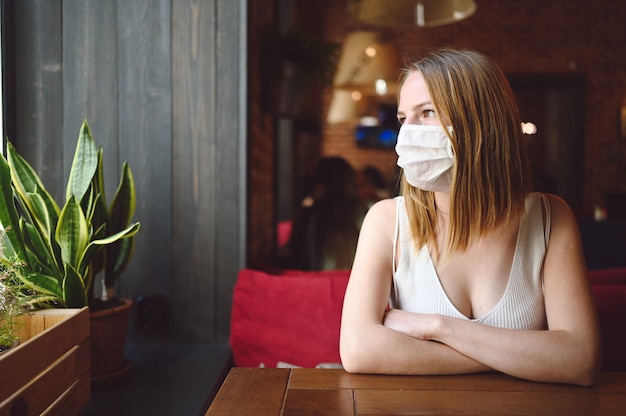 La giovane donna è seduta in un bar o in un ristorante e sta aspettando il suo ordine indossando una maschera protettiva per il viso per la prevenzione del virus dell'influenza.