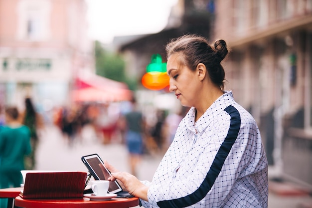 La giovane donna è seduta in un caffè di strada e lavora con il tablet Sul tavolo ci sono un taccuino e una tazza di caffè
