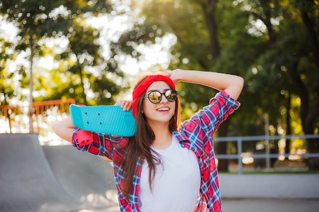 La giovane donna emozionale allegra si è vestita in vestiti alla moda mentre tiene lo skateboard in sua mano nello skatepark al giorno soleggiato luminoso