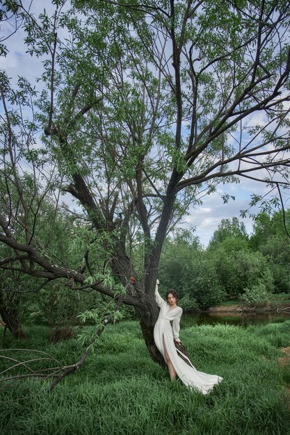 La giovane donna erotica in un vestito bianco lungo cammina in un parco di primavera vicino al fiume. Bruna sexy in posa nella natura